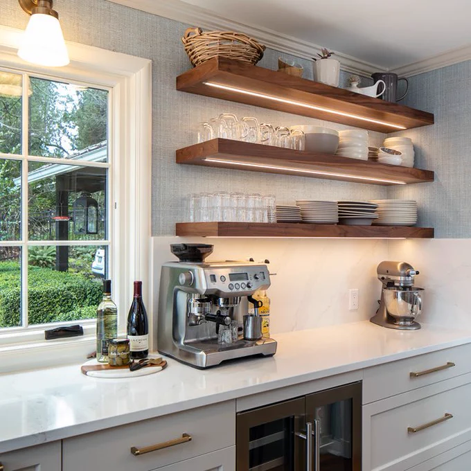 A beverage station on one wall of the kitchen with floating shelves storing glassware, a wine fridge below, and an espresso machine on the counter.  