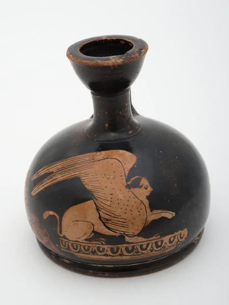 A bulbous black small vase sits on a white background. It has a thin neck and a flaring thick rim. Patches of the red body of the vessel are wearing through the glaze. A sphinx has been scratched into the black glaze, and sits on a band of ovolo / egg and dot pattern, one paw raised, with two wings.