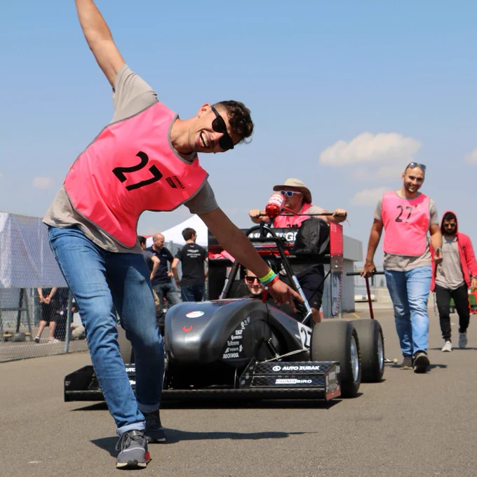 A competitor throws his arms wide and smiles for the camera as his team's black car is pushed behind him
