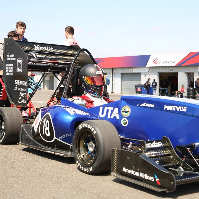 The University of Texas Arlington's team push their blue car across the silverstone paddock while the driver in the seat has his helmet down
