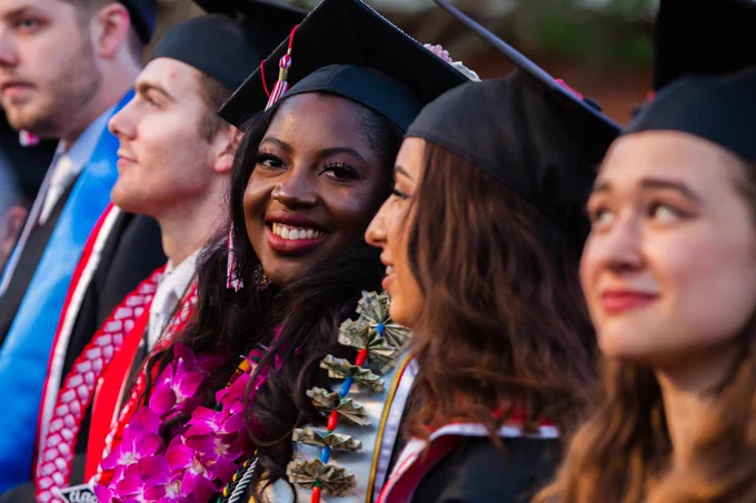 Graduate looking at camera, while other graduates sit alongside her.