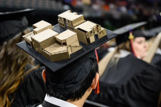 Student wearing a graduation cap with a small wooden building structure on top of it.