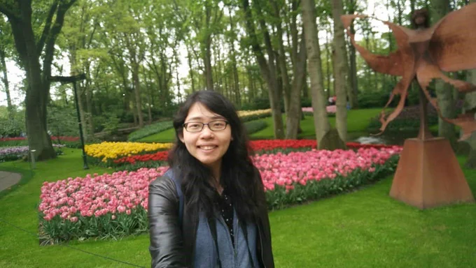 A smiling young woman faces the camera.  She’s in a park with banks of colourful tulips among grass and trees.
