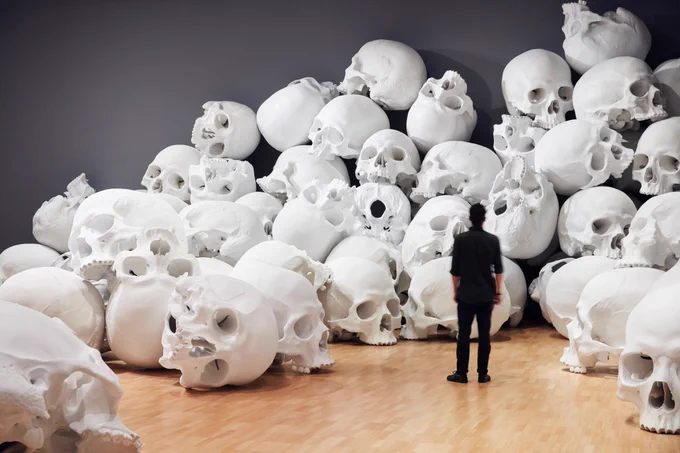 Small man in black looks at giant human skulls piled up