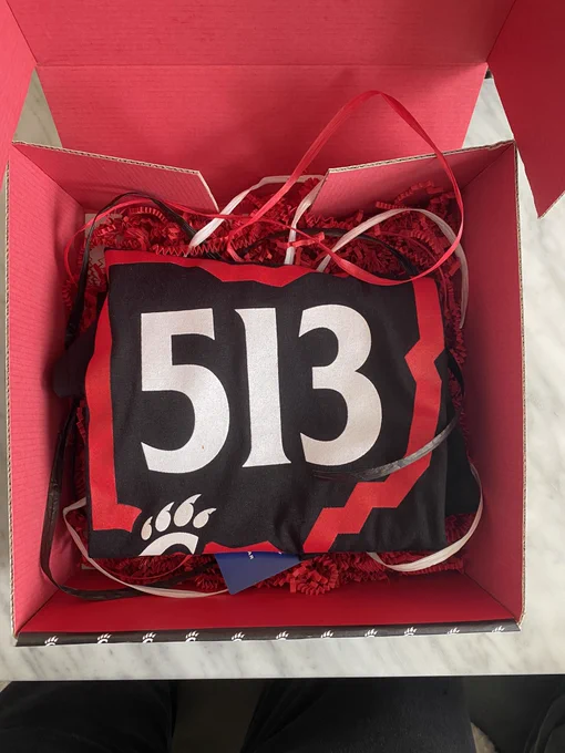 Folded black shirt with 513 on the front, folded in a box with red confetti. 