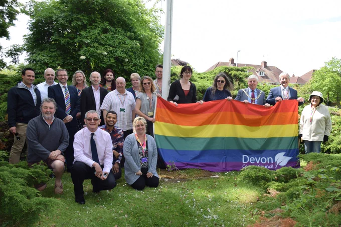 19 people stand in front of the pride flag outside County Hall, to mark the start of Exeter Pride.