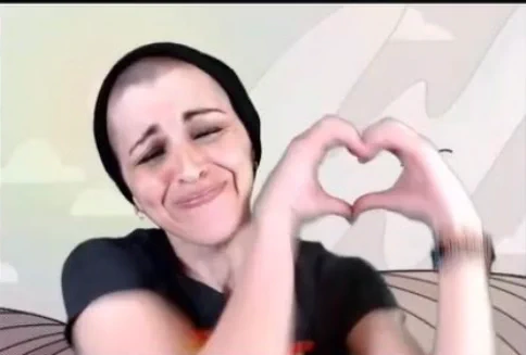 A white nonbinary person scrunches up their face and raises their hands into a heart shape