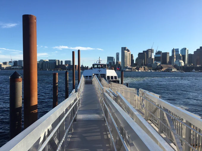 Foreground shows a ferry dock on Boston Harbor, with ferry docked bearing the MBTA logo. Downtown Boston appears in the background.
