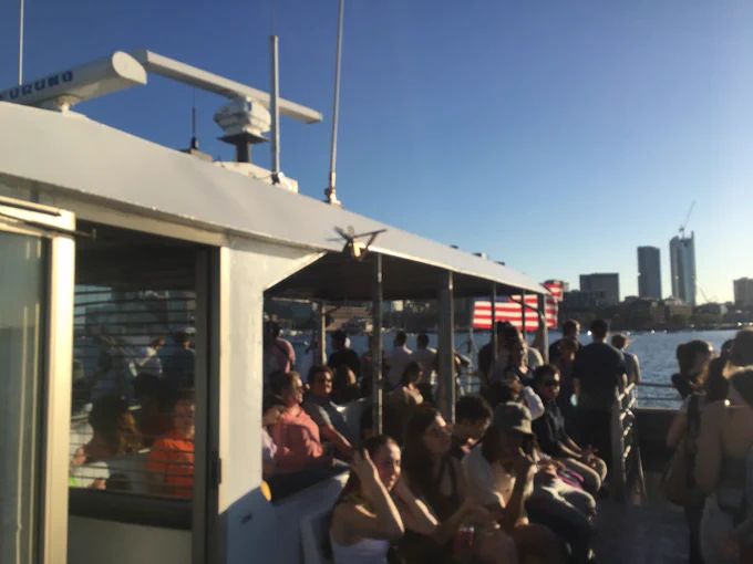 Top deck of a ferry sailing on Boston Harbor. A large throng of passengers are onboard, not hmm sitting and standing. A US flag appears at the end of the ferry. Downtown Boston high rise buildings appear in the background.