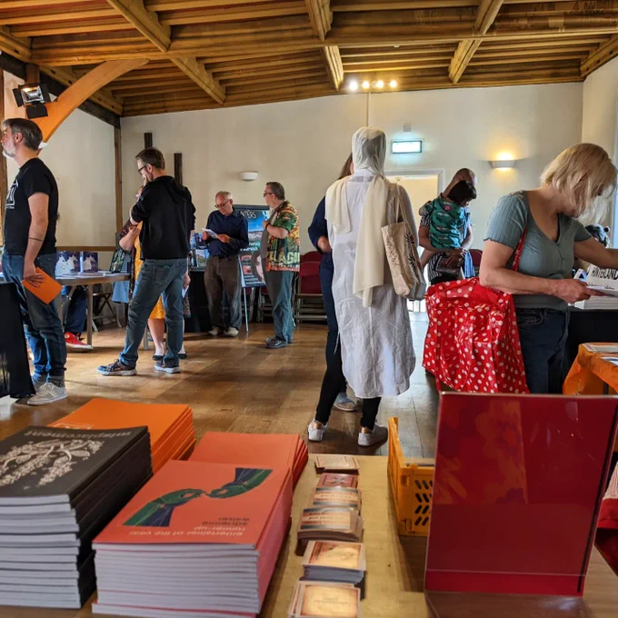 a photo taken from behind a table of books, zines and stickers at a book fair. people are talking, milling, and buying books in the background.