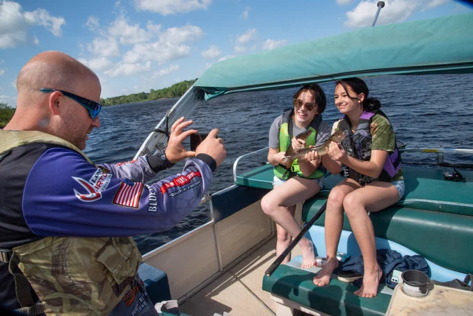 An adult takes a photo of two teens both holding northern pike up for the camera and smiling. 