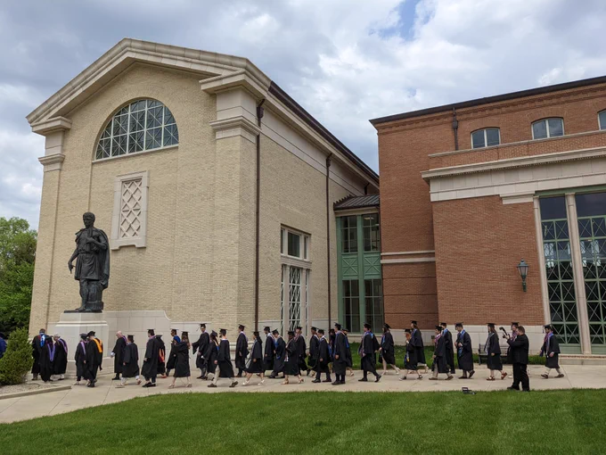 A long line of people in graduation caps and gowns walk in front of a building with a large statue of Alberti. 