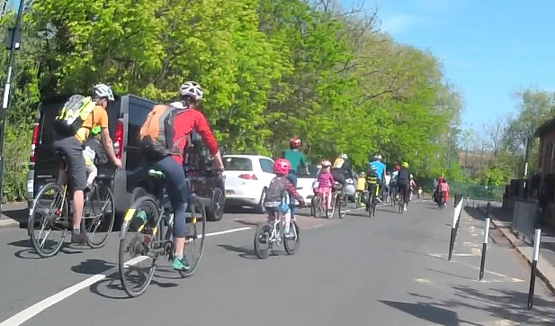 Adults and children cycling alongside parked cars next to West Jesmond Primary School.
