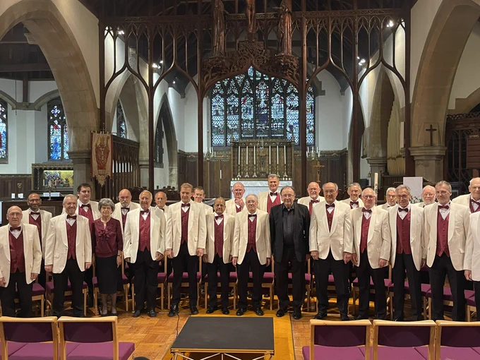 Image of about 30 men wearing cream jackets and claret waistcoat standing in front of a traditional altar, rood screen and church.  The director is wearing a black shirt and tie and the accompanies is wearing a claret shirt, both are seated either side of the centre of the front row.