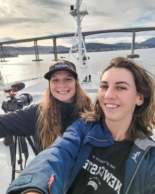 A selfie of 2 young women on a boat. They are both smiling and wearing University of Newcastle t-shirts. There is a bridge and the ocean visible in the background.