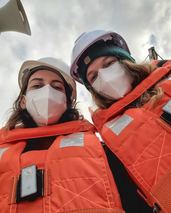 A photograph of the same 2 women as the first image. This time they are taking a selfie from a low angle looking up toward their faces. They are both wearing bright orange vests, white hard hats and white face masks.