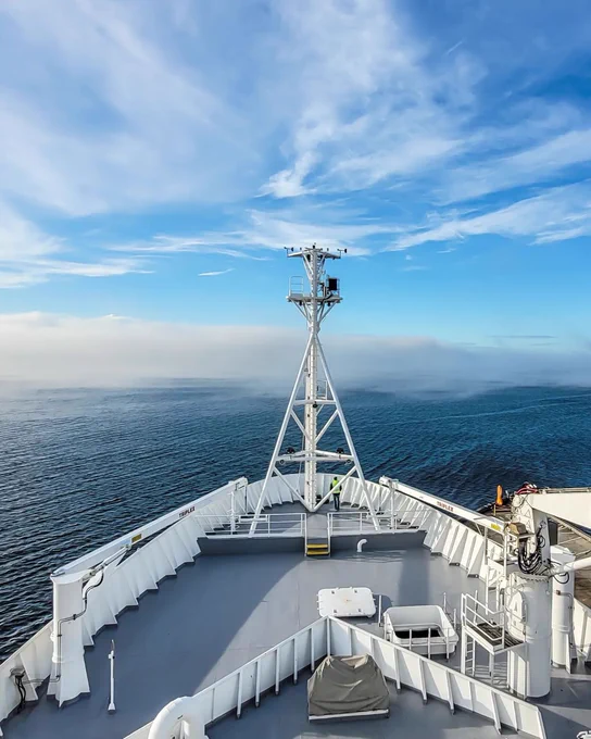 A photograph of the bow of a large boat pointing out to sea. The ocean stretches far into the distance and a low cloud hangs over the water.