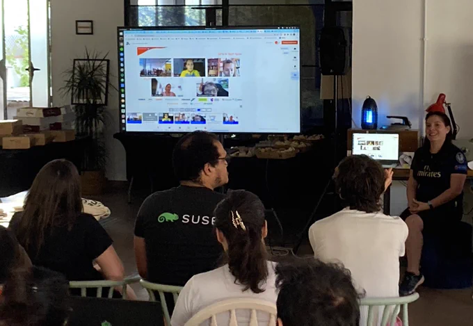 Presentación del equipo 2, presentando online desde Barcelona. De fondo la pantalla de televisión con la retransmisión de la presentación, a la derecha de la foto Francis la organizadora, y los 3 jueces del hackathon en la primera fila de sillas mirando a la presentación