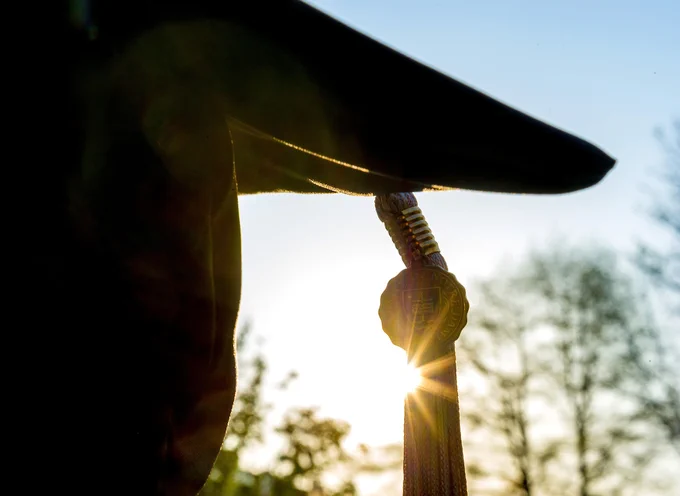 The sun flares brightly shining from behind a tassel attached to a graduation cap.