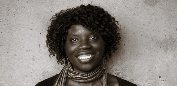 A black & white photo of Liz Ogbu, a black woman, smiling in front of a concrete wall.