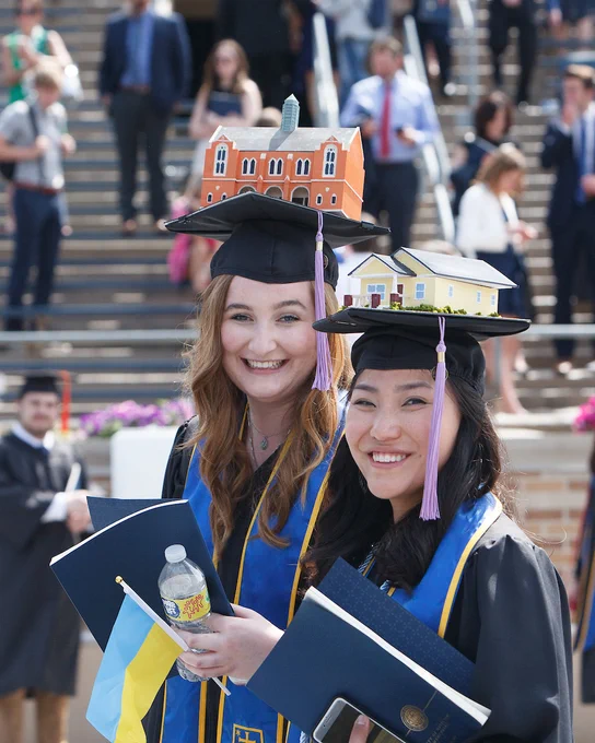 3-D structures of South Dining Hall at the University of Notre Dame, and a Habitat for Humanity house  on top of the mortarboard graduation caps of two smiling women.