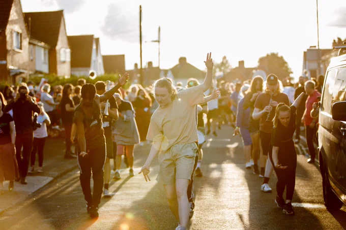 Musicians and dancers parade through a suburban street in Bristol on a sunny Summer's day. The sun is casting a golden light across the scene and the street is lined with spectators.