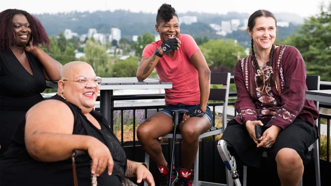 Four disabled people of color with canes and prosthetic legs laugh while chatting. They are on a rooftop deck, in chairs of various height, with greenery and city high-rises in the background.