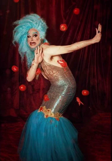 Burlesque star EmpeROAR Fabulous wearing a silver dress with a blue tulle skirt. He has blue hair and a blue beard and is putting his arms out to block flying tomatoes.