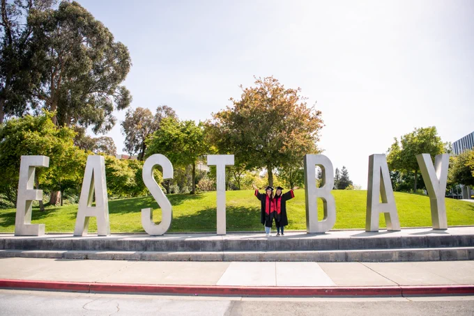 Alison and Jennifer in grad gear with arms up by East Bay sign