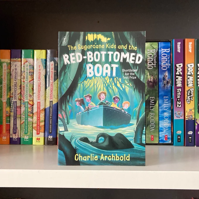 A copy of the middle-grade novel The Sugarcane Kids and the Red-Bottomed Boat, which has a bluey-green illustrated mangrove cover with a croc's head in the foreground and a boat with five kids in it, all looking in wonder and trepidation at their surroundings. The book is on a shelf of children's books including the Treehouse series by Andy Griffiths, Dog Man, and Emily Rodda's Rondo series.
