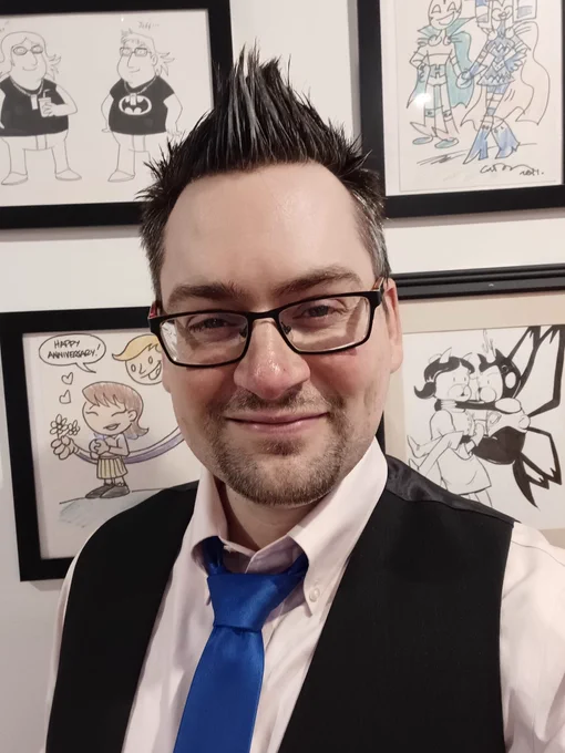 Jeff Selfie in a pink shirt, with a grey vest and blue tie. His hair is spiked up as usual, and he's very happy.

He's got a wall of stuff behind him: A drawing of Roast Beef & Molly from Achewood dressed as The Monarch & Dr. Girlfriend, Tiny Titans-style drawings of Ralph & Sue Dibny and Mister Miracle & Big Barda, and a drawing of him and Jen as Jay Sherman (their "Jay Shermansonas").