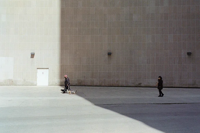 Image 1) Two people walk by a large grey wall. One of the people walks with a dog on a leash. The wall is in partial shadow.