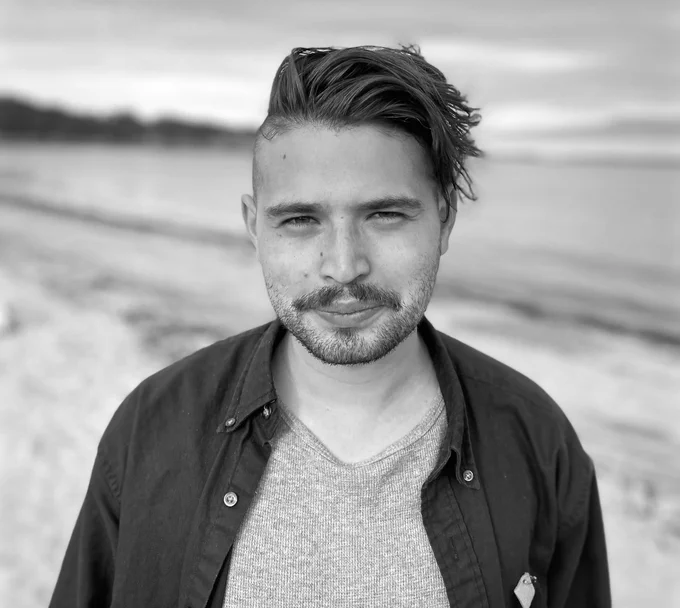 Philip geller stands on a beach with the wind blowing in his hair. They are wearing wearing an opened button shirt with a textured V-neck underneath he is looking at the camera with smiling eyes and a small grin