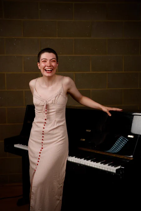 Meg, a white thin nonbinary person with shaved brown hair, is laughing as they lean on a piano. They are in a pale pink dress with red buttons down the front.