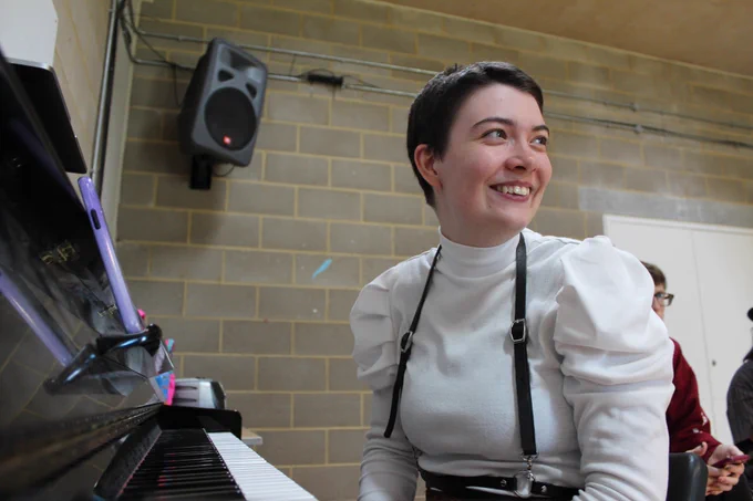 Meg, a white thin nonbinary person with shaved brown hair, is sitting at a piano looking over their shoulder. They are in a white top with big poofy sleeves.