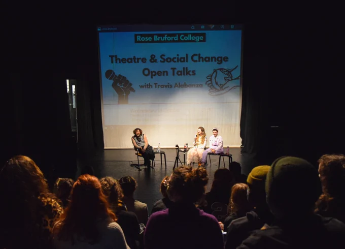 Taken from in the audience of a theatre, we can see a projection reading 'Theatre and Social Change Open Talks With Travis Alabanza'. In front of this projection sits Travis Alabanza, Nelson Nash and Meg McGrady.