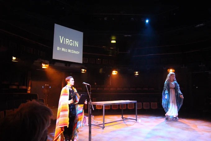 Meg, a white thin nonbinary person with shaved brown hair, is standing at a lectern. A presentation in the background reads 'Virgin by Meg McGrady'. They are dressed in a rainbow knitted blanket and a black dress with buttons down the front. The Virgin Mary is also looming in the background to the right.
