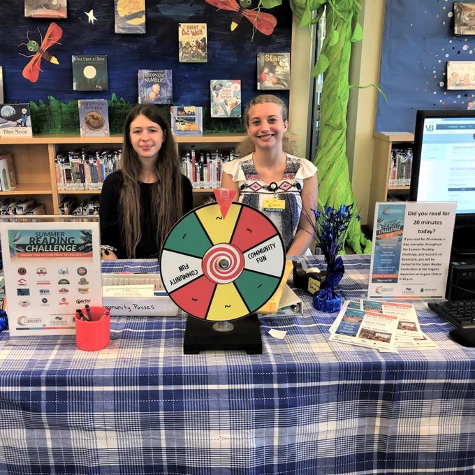 Two teen volunteers pose at a summer reading challenge prize station.