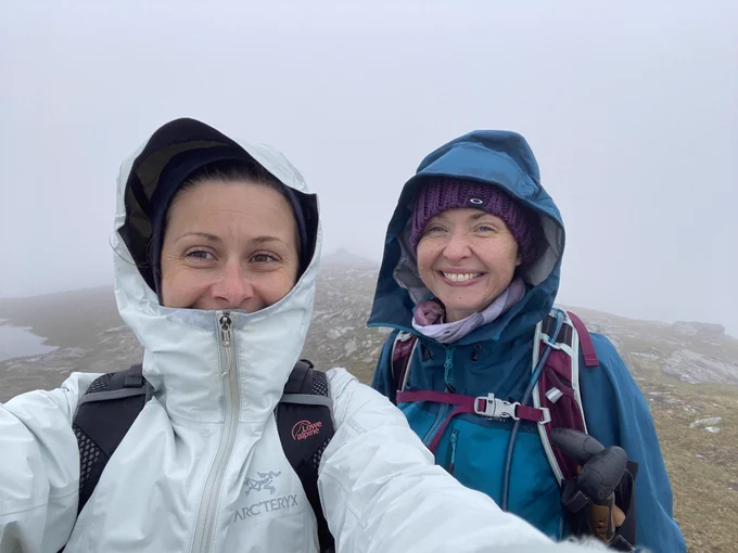 Two people are wearing waterproof jackets and smiling at the camera. A hill and fog is in the background. 