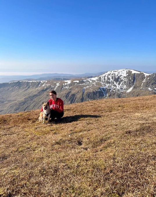 A person is kneeling down dog next to a dog. A blue sky and mountain is in the background. 
