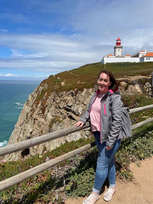 A person is smiling at the camera. In the background is a lighthouse on a hilltop. The sea is in the distance. 
