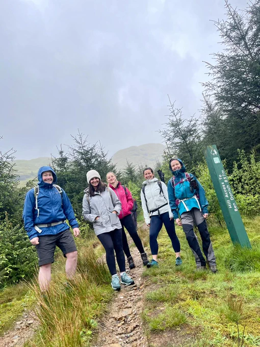 A group of people wearing jackets are smiling at the camera. They are standing on a grassy path. Tall green trees are in the distance. 