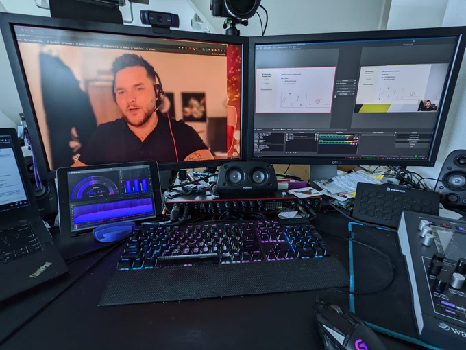 picture of my desk, there are two monitors, a laptop to the right just out of the frame. You can see the camera fees of a speaker on the left screen and OBS on the right screen. In front of the left screen there's an ipad on a stand displaying loudness meters. In the middle in front of the display there's a RGB backlit keyboard lit up in the trans pride flag colors. In front of the keyboard there is a mouse, to the right of it almost out of the frame there is a synthesizer. In front of the right screen there's a stream deck XL.