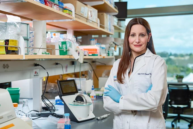 A woman stands in a lab with arms folded wearing a lab coat and gloves