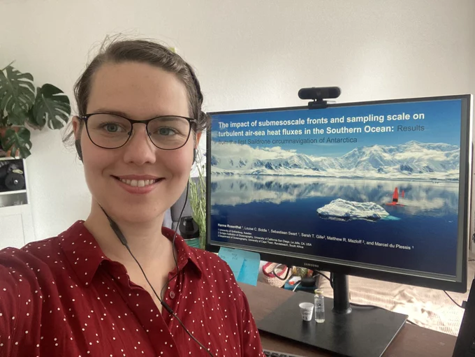 Woman in a red blouse in front of a monitor. The monitor shows an opening slide for a presentation titled “ the impact of submesoscale fronts and sampling scale on the turbulent air-sea heat flux in the Southern Ocean: Results from the first Saildrone circumnavigation of Antarctica” and a picture of an orange Saildrone passing snowcovered mountains and an iceberg on a mirror flat ocean.