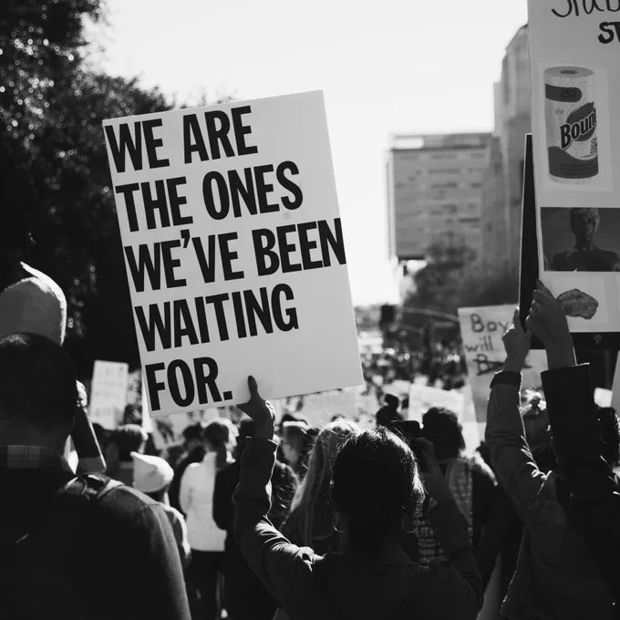 A black and white photo of people at a protest, with someone holding a sign that reads “We are the ones we’ve been waiting for”.