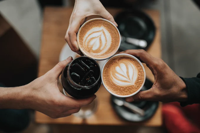 People (of which there are only 3 hands showing) holding drinks together as if saying 'cheers'. The drinks are 2 coffees, with flowers decorated in the milk, and a coke with ice.