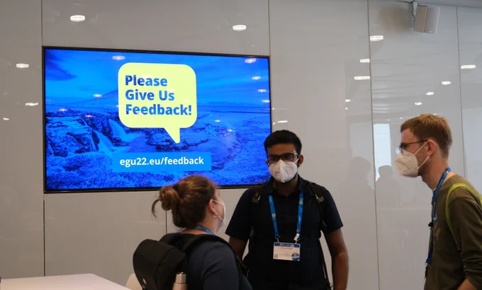 Three people wearing facemasks in front of a screen that reads: please give us feedback! egu22.eu/feedback