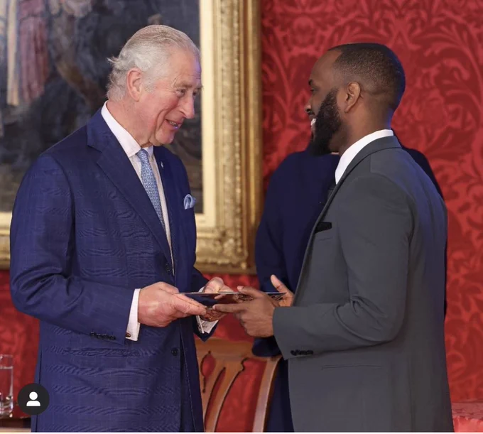 HRH The Prince of Wales handing award to young Black man Cordell Jeffers at royal event. Red background. 