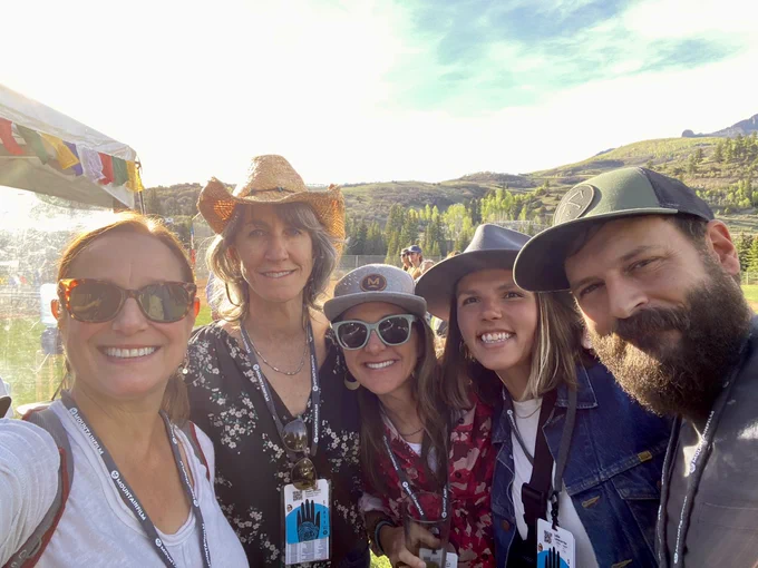 Photo of four women on the left (three in hats) and a man on the right, all huddled and smiling against the backdrop of green mountains.